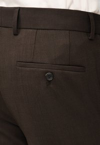 Σκούρο καφέ υφασμάτινο παντελόνι με θηλιές για ζώνη και μια πίσω τσέπη ασφαλισμένη με ένα μαύρο κουμπί, συνδυασμένο με λευκό πουκάμισο.