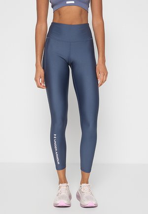 Femme portant un legging taille haute bleu marine Under Armour et des chaussures de course rose clair, debout devant un fond clair uni.