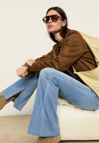 Veste bomber en daim marron, jeans évasés bleu clair avec des ourlets effilochés, bottines beiges à la cheville, lunettes de soleil marron surdimensionnées, sac à dos texturé jaune.