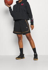Hoodie noir court avec logo rouge, short noir avec accents dorés, chaussettes noires et baskets noires. Le mannequin tient un ballon de basket.