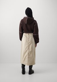 Ruskea lyhyt huppari valkoisilla saumoilla, yhdistettynä pitkään vaalean beigeen hameeseen ja mustiin polvipitkiin saappaisiin, kuvattuna takaa.