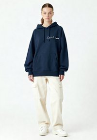 Sweat à capuche marine avec le texte blanc « Je m'en fiche », poche kangourou et capuche à cordon, associé à un pantalon cargo crème et des baskets blanches.