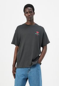 T-shirt di cotone grigio scuro con maniche corte, con un logo Adidas rosso e verde sul petto. Indossato con jeans di denim blu.
