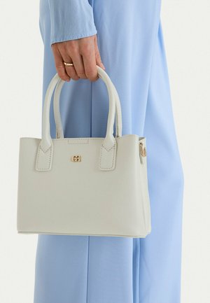 Hand mit Goldring, die eine weiße Lederhandtasche mit doppeltem Griff vor einem hellblauen Hosenhintergrund hält.