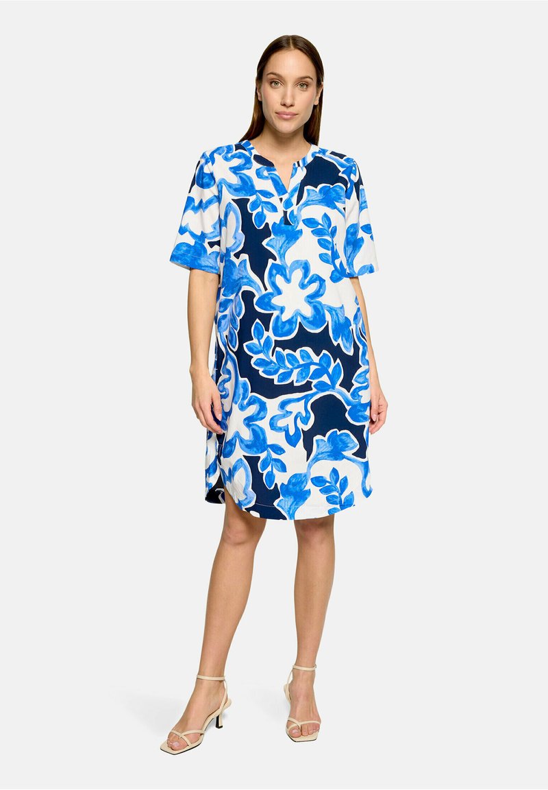 Femme debout portant une robe ample mi-longue avec des motifs floraux abstraits bleus et blancs et des sandales à talons beige à brides.