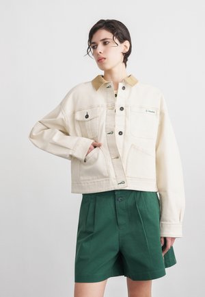 Kvinde med kort mørkt hår iført en cremefarvet jakke med beige krave og grønne højtaljede plisserede shorts, stående mod en ensfarvet baggrund.