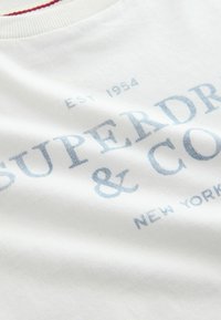 Biely bavlnený tričko s modrým potlačeným textom "SUPERDRY & CO" a "NEW YORK." Viditeľný detail rebrovaného okraja krku.