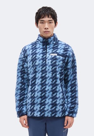 Junger Mann trägt einen blau-schwarz gemusterten Fleece-Pullover mit Reißverschluss am Kragen und Columbia-Logo auf der Brusttasche.