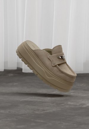 Mule en daim beige avec une semelle plateforme épaisse en caoutchouc et une boucle décorative argentée sur un sol gris lisse devant des rideaux blancs translucides.
