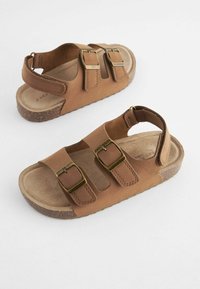 Bruna lädersandaler med breda remmar, guldfärgade spännen och en strukturerad korksula. Öppen bakdesign för andningsförmåga och komfort.