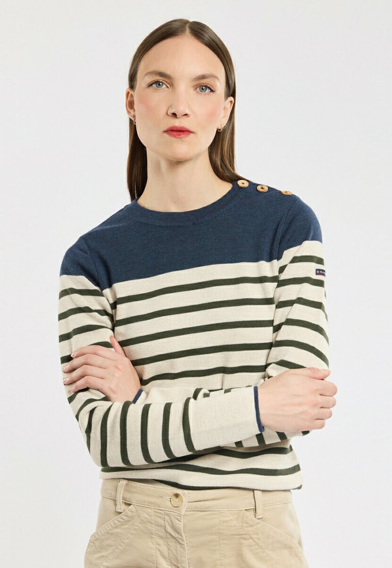 Femme aux cheveux bruns raides portant un pull rayé bleu marine et crème avec des boutons sur une épaule, croisant les bras, sur fond blanc.
