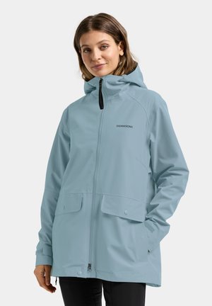 Woman wearing a light blue hooded rain jacket with front zipper and flap pockets, standing against a plain background.