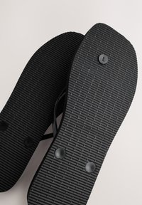 Tongs noires avec semelles texturées et attaches de boutons visibles en dessous, placées en chevauchement sur une surface claire.