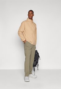 Veste beige clair avec un col haut, associée à un pantalon cargo vert olive. Le modèle porte un sac à dos noir et des baskets blanches.