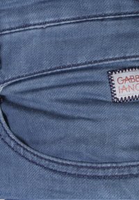Tessuto denim con una texture liscia in una tonalità di blu medio. Presenta una tasca posteriore e un'etichetta cucita con filati rossi e bianchi.