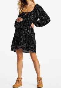 Žena v černých dlouhých šatech s bílými puntíky, v hnědých šněrovacích kotníkových botách, stojí s jednou rukou na boku.