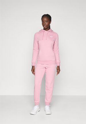 Lys pink hoodie og sæt med sweatpants lavet af blødt stof, med en kengurulomme og broderet logo, parret med hvide sneakers.