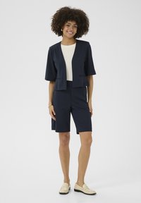Blazer blu navy con maniche corte e apertura frontale, abbinato a pantaloni corti al ginocchio dello stesso colore. L'outfit presenta un top di colore crema sotto.