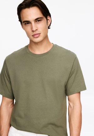 Hombre con cabello oscuro y corto, vistiendo una camiseta de manga corta lisa de color oliva, de pie contra un fondo blanco, mirando ligeramente hacia adelante.