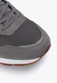 Zapatilla deportiva gris que presenta una combinación de malla texturizada y materiales de ante, con una entresuela blanca y una suela de goma marrón.
