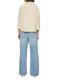 Beżowa kurtka bez kołnierza z rękawami o długości trzech czwartek; w połączeniu z jasnoniebieskimi, szerokimi dżinsami z tylnymi kieszeniami.