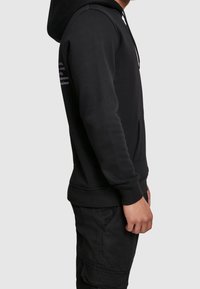 Felpa nera con cappuccio regolabile, tasca frontale e polsini a coste. Presenta una scritta stampata sulla manica sinistra. Il materiale sembra morbido e strutturato.