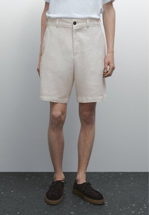 Beige linen shorts with a straight cut, featuring a front button closure, two side pockets, and a smooth texture. Brown suede shoes complete the look.