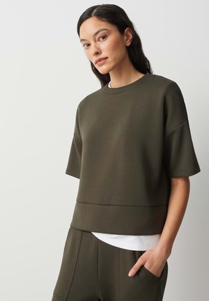 Donna con capelli scuri che indossa una maglietta verde oliva a maniche corte e pantaloni abbinati, in piedi con una mano in tasca su uno sfondo semplice.