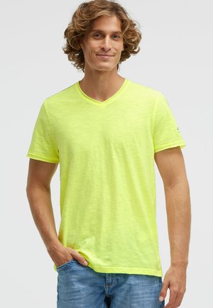 Junger Mann mit lockigem Haar, der ein leuchtend gelbes T-Shirt mit V-Ausschnitt und blaue Jeans trägt, lächelt und hat eine Hand in der Tasche.