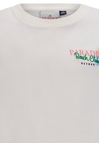 T-shirt bianco in cotone con scollatura tonda. La parte frontale presenta un testo ricamato colorato: "PARADISE Beach Club RETOUR." Texture liscia.
