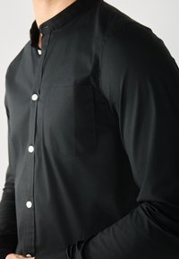 Svart knapp-up skjorta i slät tyg, med bandkrage, långa ärmar och en ficka på vänster bröst. Vita knappar kontrasterar mot tyget.