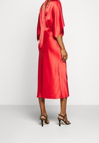 Robe midi en satin rouge avec un nœud dans le dos, des manches kimono et une fente sur le côté, associée à des talons noirs avec lanières et un bout pointu.