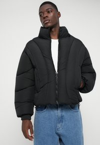Mladý muž v čiernej oversized puffrovacej bunde, pod ktorou má bielu košeľu a modré džínsy, stojí s jednou rukou v vrecku.