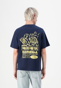 Person with short blonde hair wearing a navy blue shirt with yellow turtle and electronics store graphics on the back, paired with light jeans.