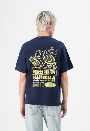 Persona con pelo corto rubio que lleva una camiseta azul marino con un gráfico de una tortuga amarilla y de una tienda de electrónica en la espalda, combinada con jeans claros.