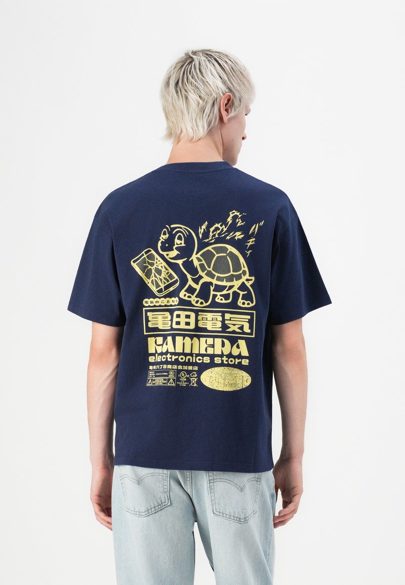 Person with short blonde hair wearing a navy blue shirt with yellow turtle and electronics store graphics on the back, paired with light jeans.