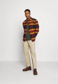 Camisa de franela a cuadros en negro, rojo y amarillo, llevada sobre una camiseta verde, con pantalones beige y botas de tobillo burdeos.