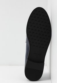 Semelle en caoutchouc noir avec grip texturé, portant un logo en relief et des indications de taille. Cuir supérieur lisse en bleu marine visible sur le côté. Design épuré.