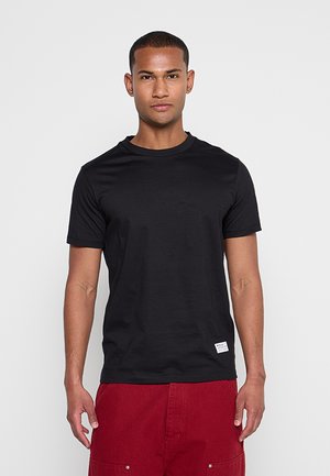 Mann mit kurzen Haaren, der ein schwarzes T-Shirt mit Rundhalsausschnitt und rote Hose trägt, steht vor einem schlichten hellen Hintergrund.