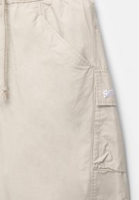 Pantaloni corti beige realizzati in tessuto leggero. Presentano un cintura regolabile con lacci, tasche laterali e una patch con il logo sul lato. Tessuto morbido.