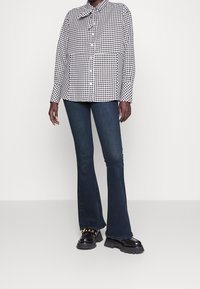 Persona que lleva una camisa de cuadros en blanco y negro, pantalones vaqueros de campana de color azul oscuro y zapatos de charol negros con ojales dorados.