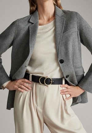 Kvinde iført en grå blazer over en beige top, der er stoppet ned i cremefarvede bukser med høj talje og sort bælte med dobbelte gyldne ringe.