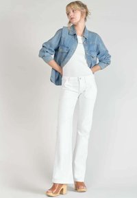 Un jean blanc évasé associé à un haut blanc ajusté et à une veste en denim bleu clair. Des sabots en bois brun à talons complètent l'ensemble.