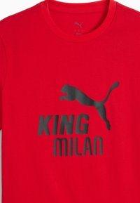 Rød bomulds T-shirt med et stort sort Puma-logo og teksten "KING MILAN" i fede sorte bogstaver, rund halsudskæring og korte ærmer.