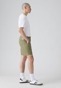 Șorturi pentru bărbați de culoare verde măslinie, fabricate dintr-un material durabil, dotate cu buzunare laterale, un buzunar pe spate cu fermoar și o croială regulată, asortate cu un tricou alb.