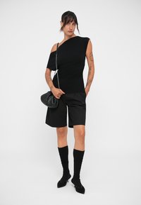 Top corto nero a maniche corte con scollo asimmetrico, abbinato a pantaloni corti neri su misura, calze nere fino al ginocchio e una piccola borsa nera.