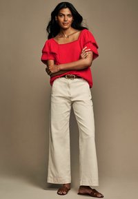 Blusa rossa con scollatura quadrata e maniche a volant, abbinata a pantaloni larghi color crema. Una cintura marrone e sandali piatti completano l'outfit. Tessuto liscio.