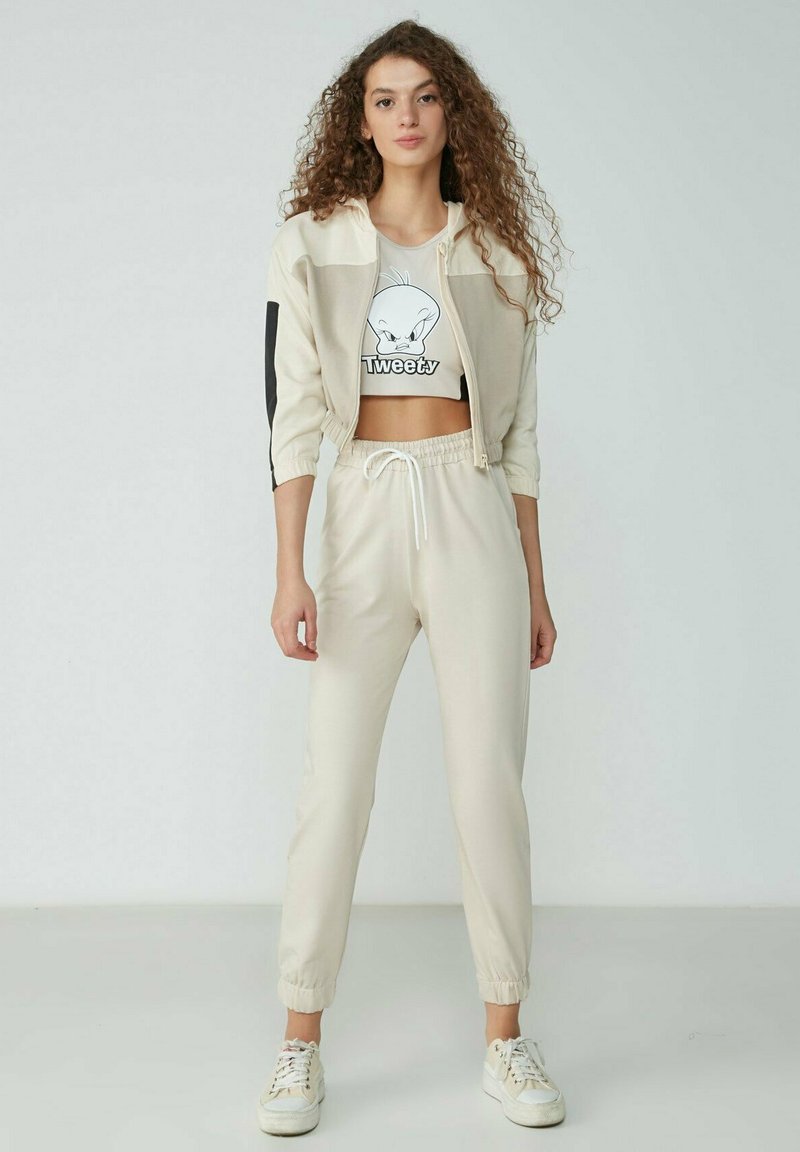 Beige cropped Jacke mit schwarzen Akzenten, kombiniert mit beigen Jogginghosen. Das Oberteil hat einen Cartoon-Charakter-Druck. Weiße Sneaker runden das Outfit ab.