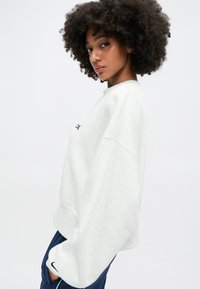 Svetlošedý oversized sweatshirt s okrúhlym výstrihom a zníženými ramenami, s malým čiernym logom na prednej strane a rebrovanými manžetami.