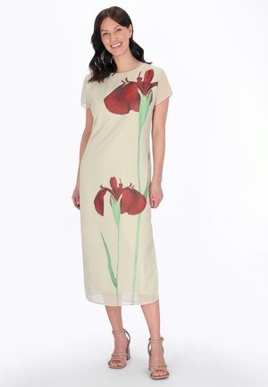 Kobieta w beżowej sukience midi z dużym czerwono-zielonym kwiatowym wzorem, uśmiechająca się i stojąca z luźno opuszczonymi rękami, w beżowych sandałach na obcasie.